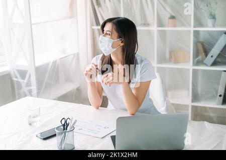 Pandemic furlough. Office relocation. Unemployment crisis. Pensive sad female employee in protective face mask thinking on new project at light workpl Stock Photo