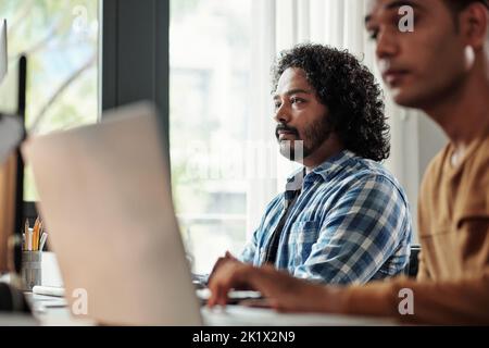 Pensive Indian programmer with curly hair looking at computer screen Stock Photo