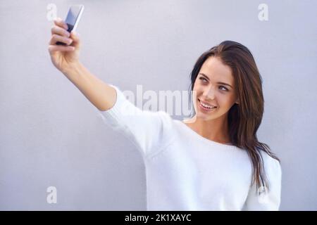 Shes selfie savvy. an attractive young woman standing against a gray background and taking a self portrait. Stock Photo