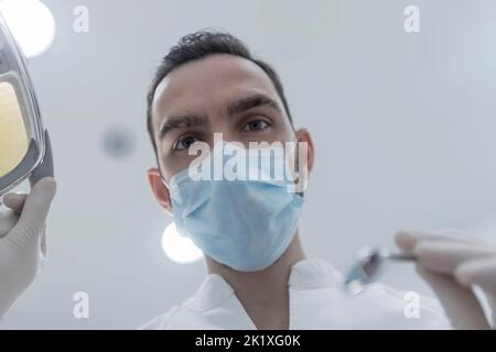 Low angle view of young dentist holding dental tools, ready for check up, professional stomatologist doctor wearing medical mask checking patient's te Stock Photo