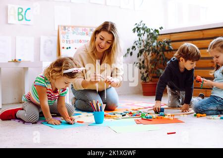 Smiling female teacher play with children, make craft, colorful handmade arts in development center for special kids. Stock Photo