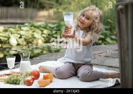 Little blonde girl present, holding milk glass in hand on picnic. Having healthy, fresh organic lunch in park near lake Stock Photo