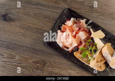 Cheese, meat plate with ciabatta bread for aperitif, appetizer on black plate on wood table in cafe. Delicatessen order Stock Photo