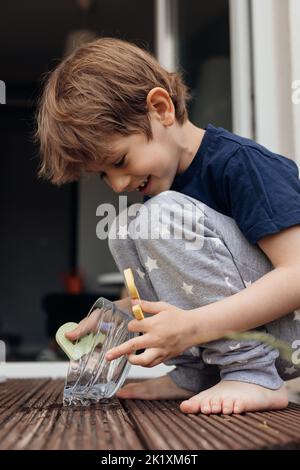 Vertical photo of little barefoot fair haired smiling boy washing and pouring clean water in animal bowl for his dog  Stock Photo