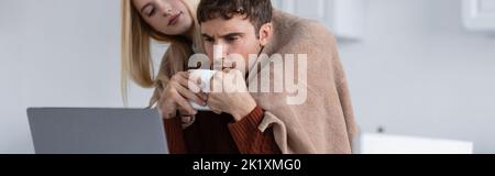 blonde woman holding blanket near boyfriend with cup working remotely from home, banner,stock image Stock Photo