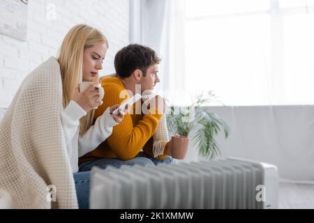 young woman with cup of tea using smartphone near boyfriend covered in blanket while sitting near radiator heater,stock image Stock Photo