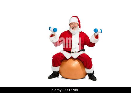 Portrait of senior man in image of Santa Claus sitting on exercise ball and doing exercises with dumbbells isolated over white background Stock Photo