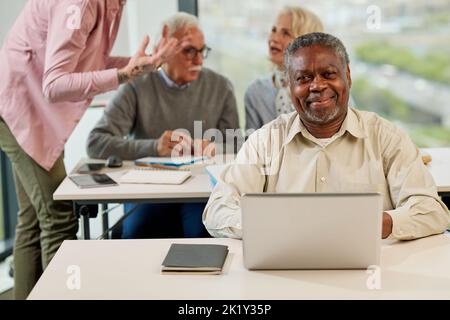 A senior african student smiling at the camera while group of multicultural senior students is discussing a programming project with an educator in th Stock Photo