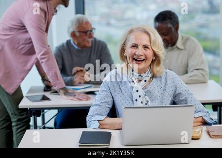 A happy female senior student sits in a classroom and does an assignment on the laptop while smiling at the camera. Stock Photo
