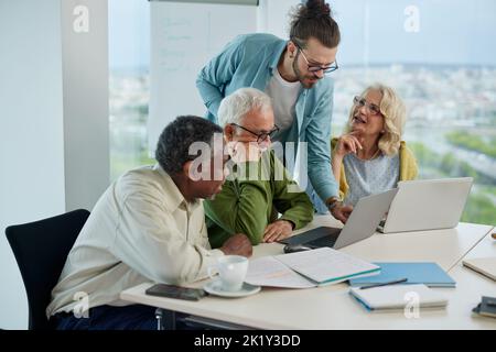 A small group of multicultural senior students is listening to the teacher's explanation and looking at the laptop. Stock Photo