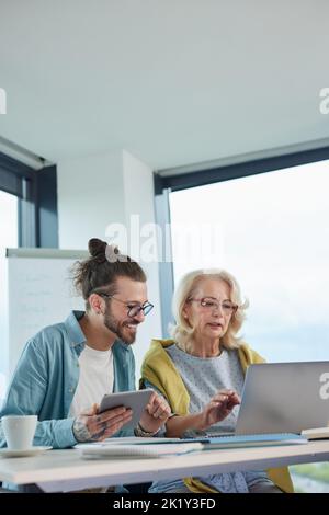 A young hipster educator is teaching a senior female student how to design a website in the classroom. Stock Photo