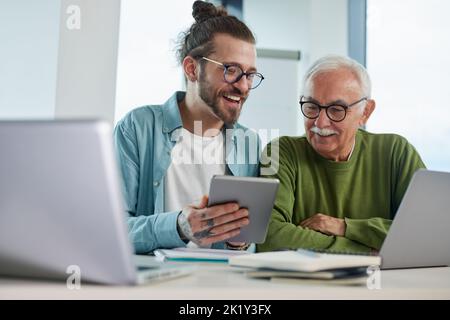 Young educator showing senior student how to solve a problem on project on his tablet while the student typing on a laptop. Stock Photo