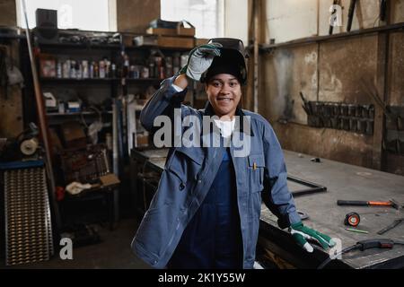 Waist up portrait of multiethnic female welder smiling at camera in industrial factory workshop, copy space Stock Photo