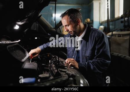 Side view portrait of mature mechanic fixing car engine in dark garage shop Stock Photo