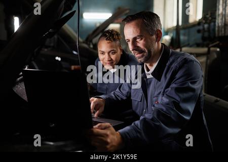 Portrait of two smiling mechanics repairing truck and using laptop in garage with accent light Stock Photo