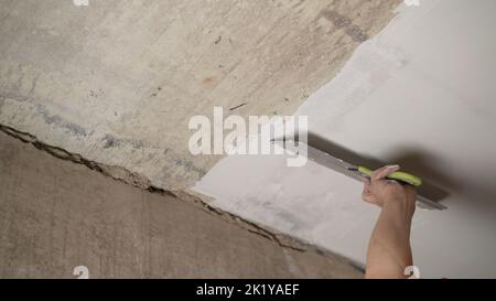 Hand with a putty knife and putty, apply mastic to the ceiling to level the surface. The process of applying mastic with a spatula to the surface to Stock Photo