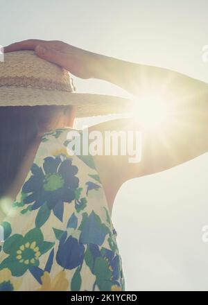 Close rear view of woman in a summer dress holding on to her straw hat on her head with intense sunburst breaking through the bent arm crease. Stock Photo