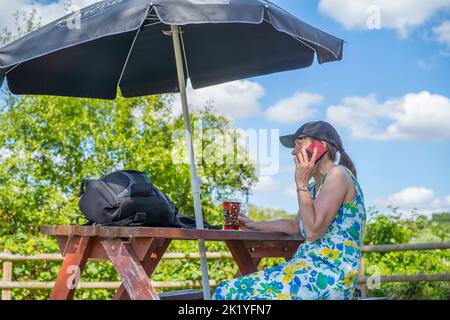 Side view of a woman sat drinking in a pub garden on a sunny day while talking on her mobile phone. The woman shelters under a sun garden brolly to pr Stock Photo