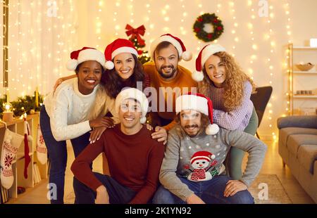 Portrait of cheerful young friends in Santa hats posing in cozy living room with Christmas decor.