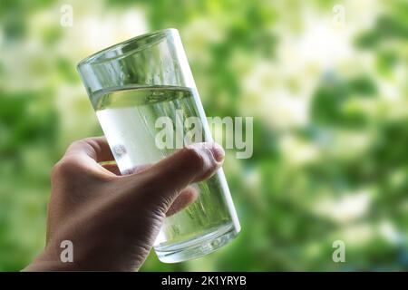 A glass of water in a hand close-up on a natural green background outdoors.. Stock Photo