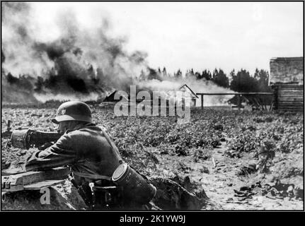 WW2 OPERATION BARBAROSSA  Counter-partisan action by Whermacht Nazi German soldiers on the Eastern Front. In the foreground, a German soldier in a trench position, firing an MG-34 machine gun with a drum magazine. A burning village in the background. 1941 Stock Photo