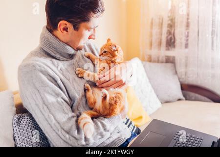 Man holding cat working in internet from home using laptop. Young freelancer playing with pet sitting on couch. Remote job Stock Photo