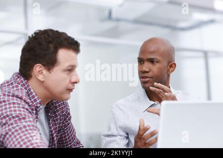 Running their ideas by each other. A young designer trying to explain something to his colleague as they sit by a desk with a laptop. Stock Photo