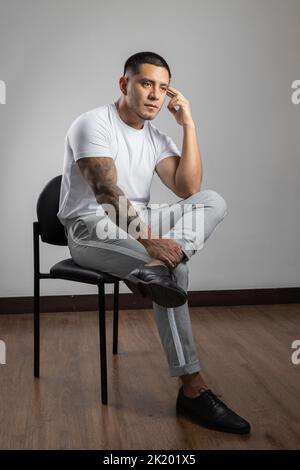 thinking sitting on a chair a young latin man with short hair and beard, wears a white t-shirt and pants in studio, model lifestyle Stock Photo