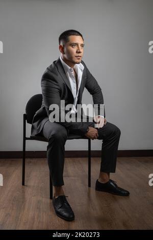 elegant latin young man with short hair and beard, business clothes, suit, shirt and tie, fashion and lifestyle in studio sitting on a chair posing Stock Photo