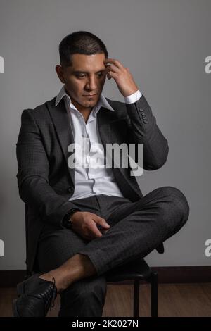 sitting on a chair a worried young latin man with short hair, business clothes, suit and shirt, fashion and lifestyle in studio Stock Photo