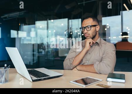 Thinking Hindu businessman in glasses, male programmer developer working inside office of modern product IT company, entrepreneur at work with laptop. Stock Photo