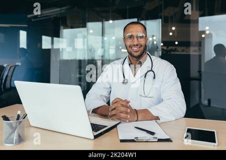 Portrait of successful male doctor, hispanic smiling and looking at camera, doctor in medical gown working inside modern clinic office, using laptop for work. Stock Photo
