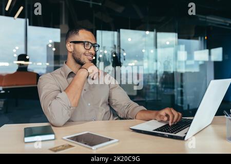 Happy and successful Indian man working in modern office building, programmer in glasses using laptop to write code, businessman in shirt smiling and happy . Stock Photo
