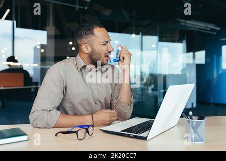 Sick office worker with asthma having trouble breathing, man using inhaler to relieve breathing, hispanic man working inside modern office building, mixed race man with laptop Stock Photo