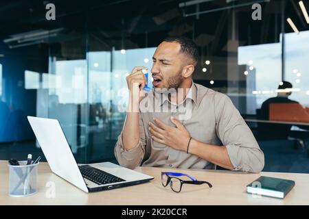 Sick office worker with asthma having trouble breathing, man using inhaler to relieve breathing, hispanic man working inside modern office building, mixed race man with laptop Stock Photo