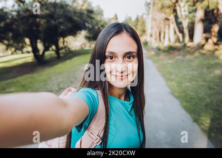 Smiling young girl wearing a turquoise t shirt and headphones, taking a POV selfie with her mobile in a natural space Stock Photo