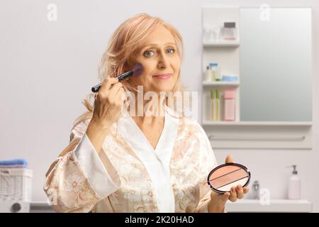 Mature woman in a bathrobe applying make up using a brush inside a bathroom Stock Photo