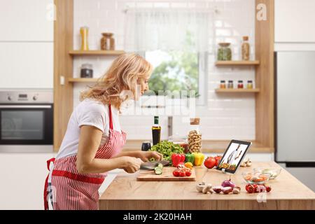 Mature woman cutting vegetables and watching a video on a tablet in a kitchen Stock Photo