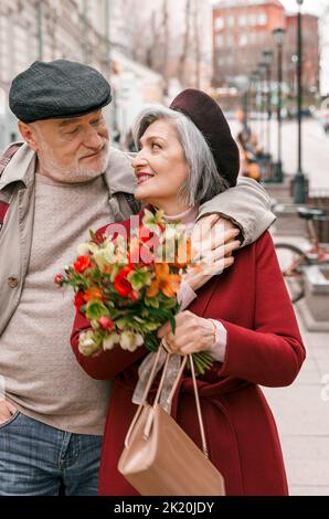 Elderly senior love couple. Old retired man woman together on romantic date.Aged husband wife walking on city street with flowers.Stylish elder huggin Stock Photo