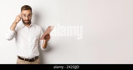 Displeased employer scolding staff while checking report on digital tablet, pointing at head and looking disappointed, standing over white background Stock Photo