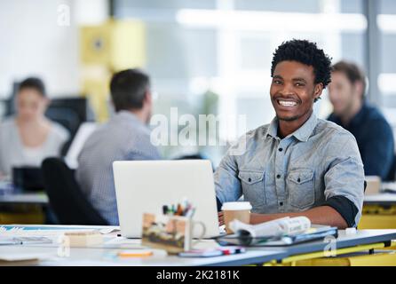 Its been a fun ride getting to where I am. Portrait of a designer sitting at his desk working on a laptop with colleagues in the background. Stock Photo