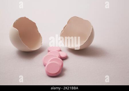 Close-up of cracked opened egg shells with pink medicine pills on light gray background. Stock Photo
