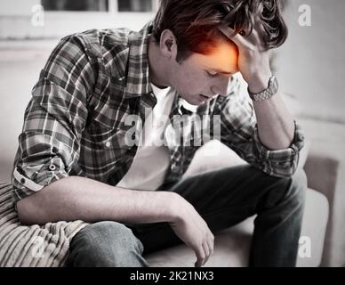This headache is killing me. A young man sitting on a couch with his hand in his hair as if in pain. Stock Photo