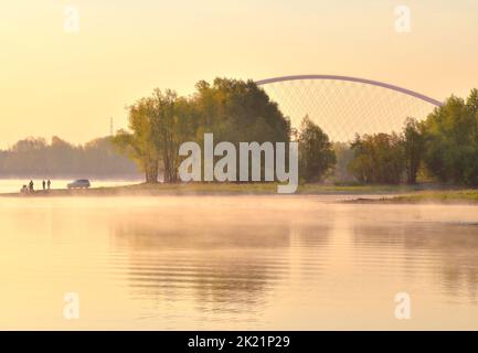 Morning on the banks of the Ob. Trees and silhouettes of fishermen are reflected in the water of the river in the morning fog, an arched bridge in the Stock Photo