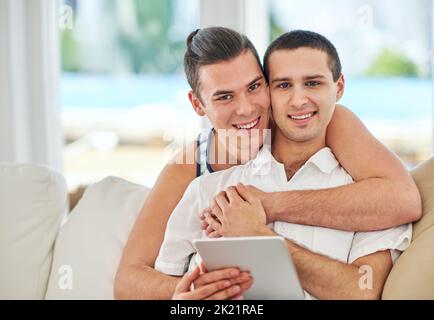 All smiles and love. Portrait of an affectionate gay couple using a digital tablet while relaxing on the sofa at home. Stock Photo