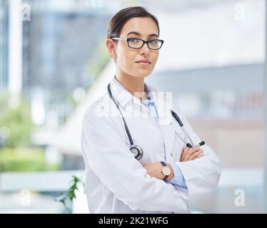 Portrait of a doctor with arms crossed in her consultation office with glasses and a stethoscope. Woman healthcare professional standing in a room in Stock Photo