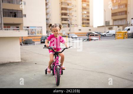 Cute little smiling girl riding bike bicycle in city on parking sunny summer day. Active family leisure with kids. Little caucasian girl 5 years old have fun with pink child bicycle Stock Photo