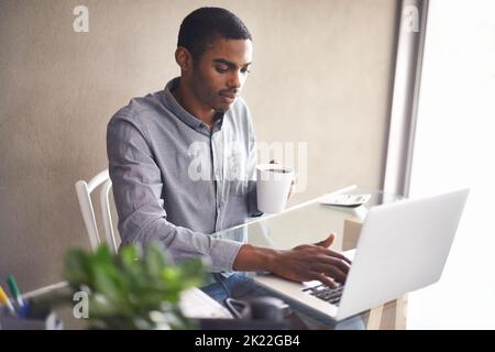 He gets a lot done in this office. a handsome young man drinking coffee while working on his laptop at home. Stock Photo