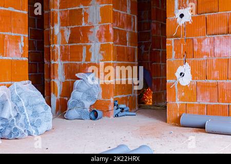 Stack of arranged parts of new grey plumbing sewer pipes elbows placed in mesh sacks, exposed electrical sockets on the wall with blocks of building u Stock Photo