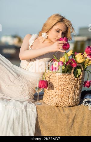 Charming woman in romantic dress smell bouquet of pink and yellow tulips in wicker basket on roof terrace. Elegant lady relax with glass of champagne Stock Photo